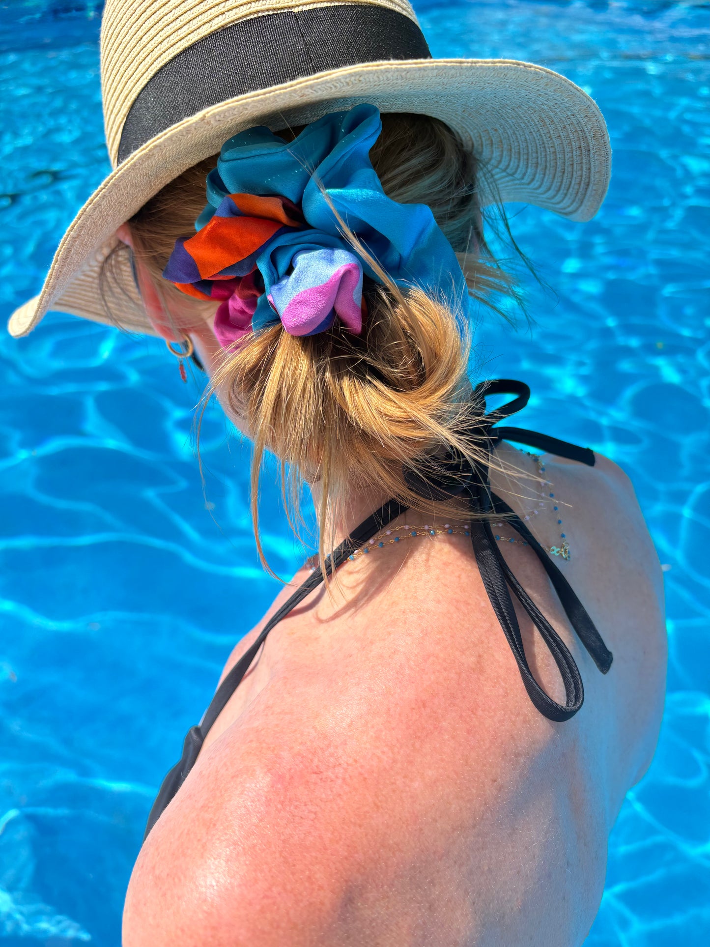 Person wearing a wide-brimmed hat with colorful scrunchie duo in their hair by a pool