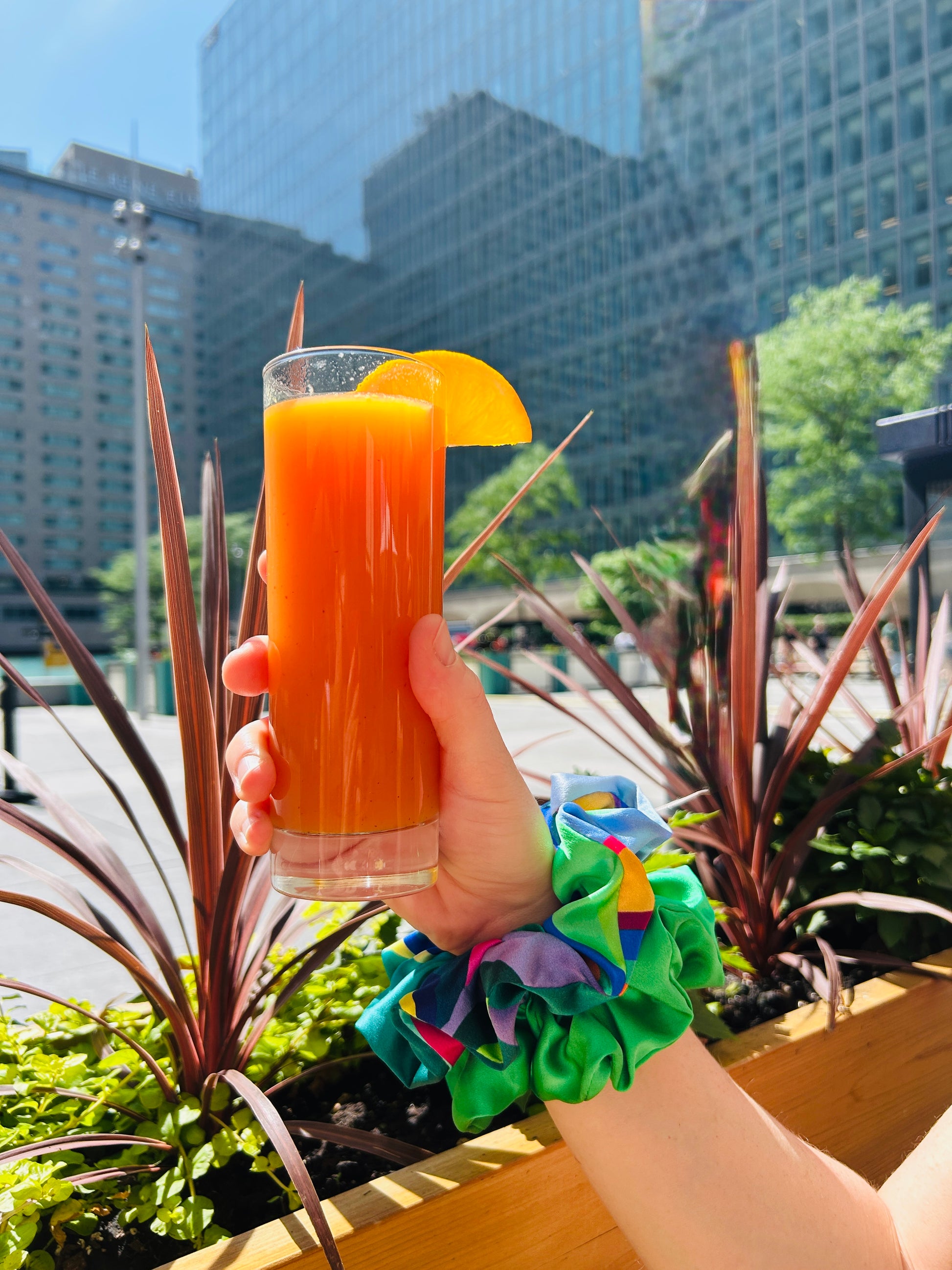 Hand holding a glass of orange juice with a scrunchie duo as wristband in an urban setting