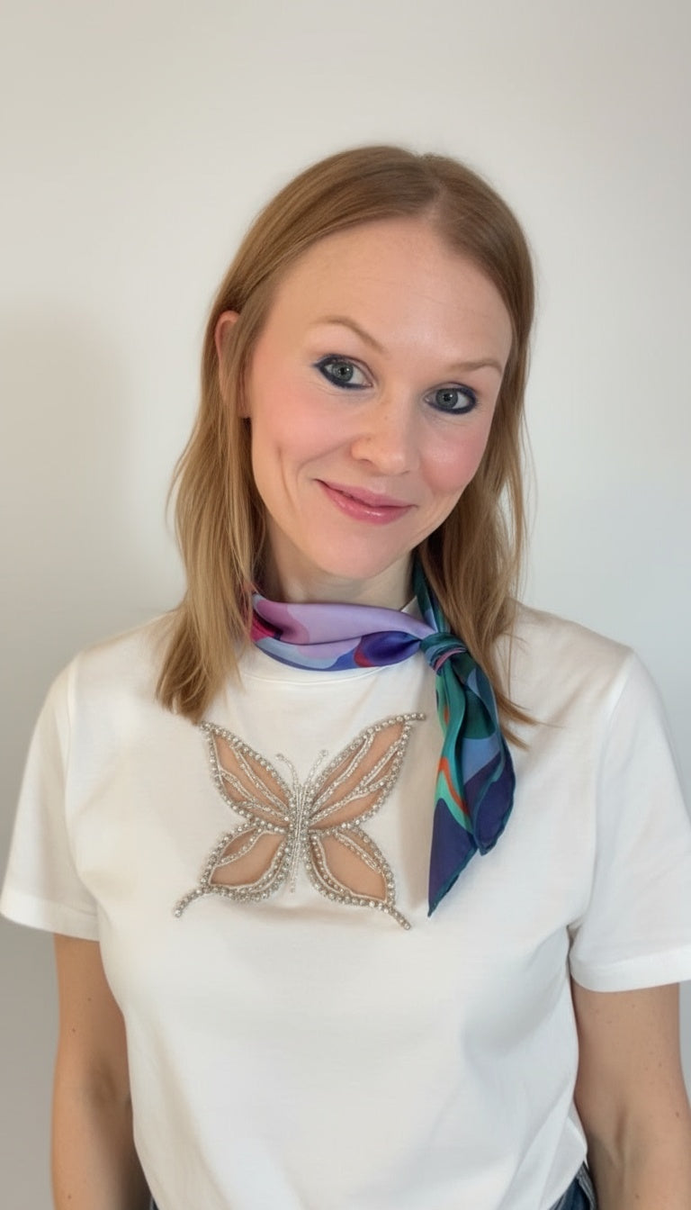 Woman wearing a white t-shirt with a decorative butterfly design and a colorful scarf, against a plain background.